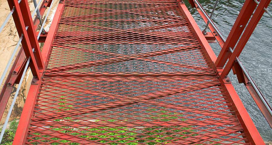 Red coated expanded metal walkway bridge with steel wire rope handrails over water